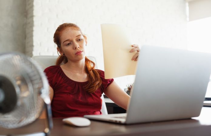 Eine Frau am Schreibtisch wedelt mit Papier Luft ins Gesicht