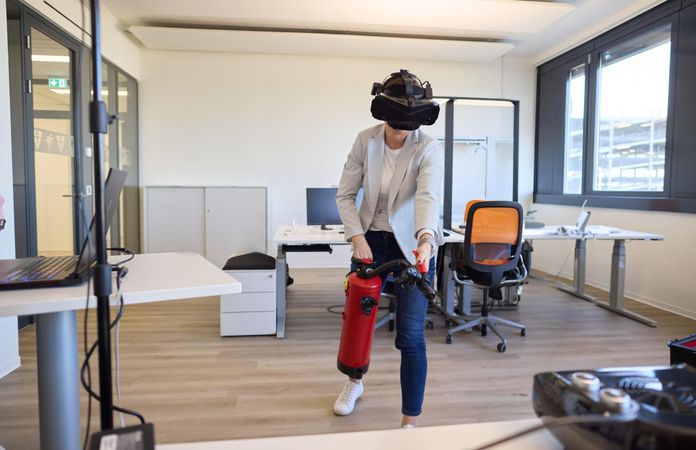 Person mit VR-Headset benutzt einen Feuerlöscher in einem modernen Büro und simuliert dabei ein virtuelles Erlebnis.