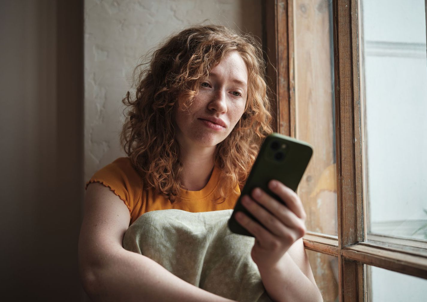 Eine Frau mit lockigem Haar und einem gelben Shirt sitzt am Fenster und schaut mit nachdenklichem Gesichtsausdruck auf ihr Smartphone.