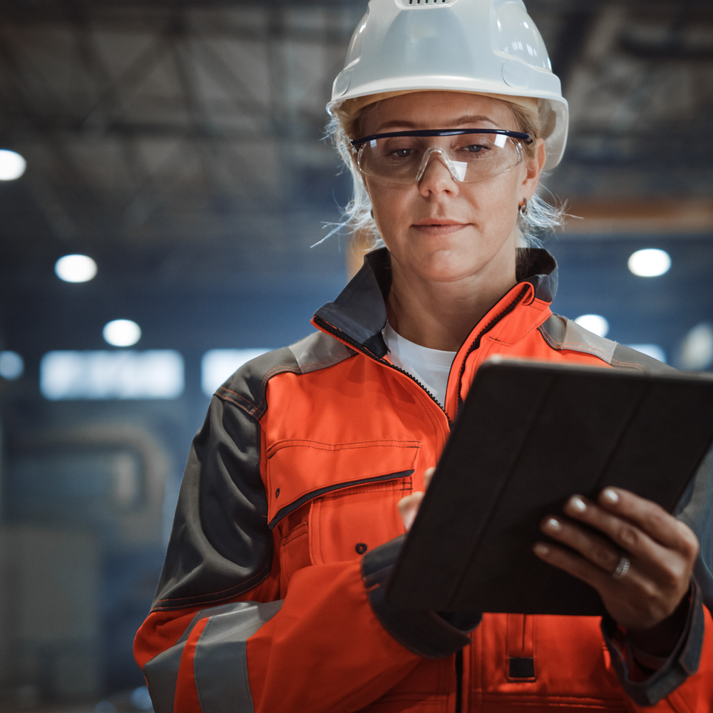 Eine Fachkraft für Arbeitssicherheit mit Helm und Schutzbrille nutzt ein Tablet in einer gut beleuchteten Produktionshalle.