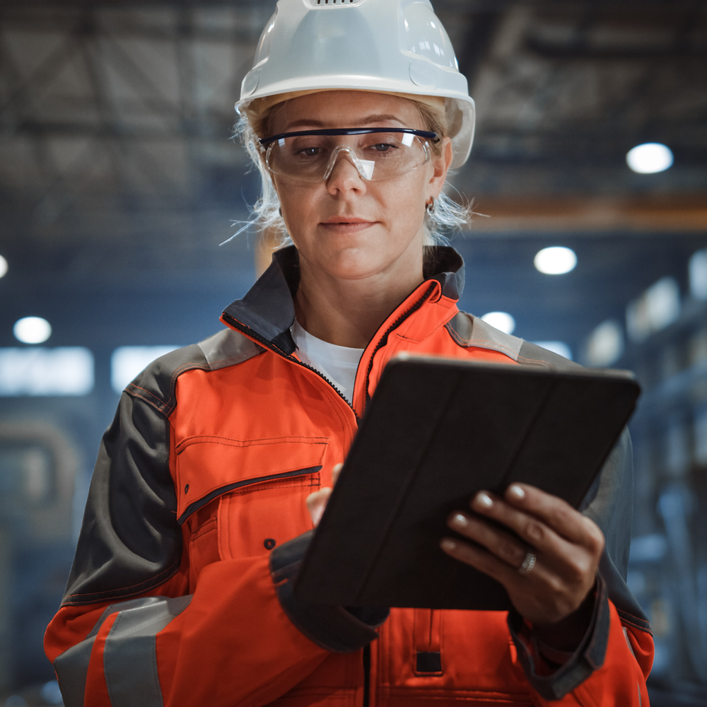 Eine Fachkraft für Arbeitssicherheit mit Helm und Schutzbrille nutzt ein Tablet in einer gut beleuchteten Produktionshalle.