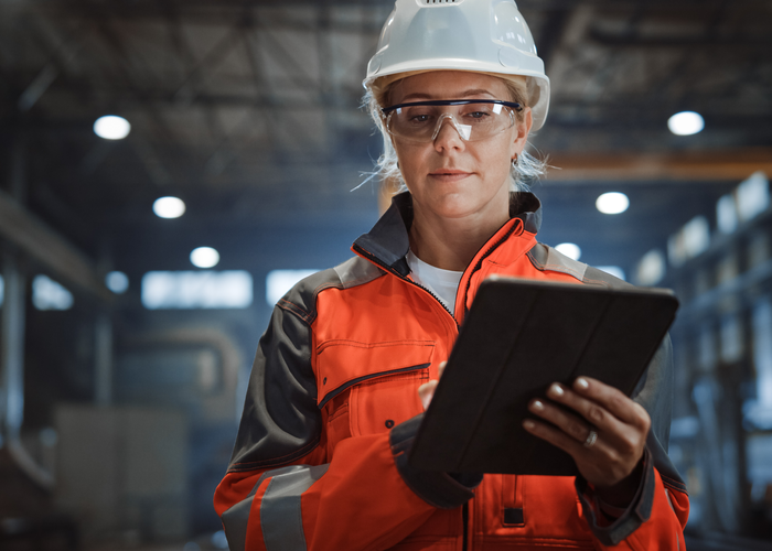 Eine Fachkraft für Arbeitssicherheit mit Helm und Schutzbrille nutzt ein Tablet in einer gut beleuchteten Produktionshalle.