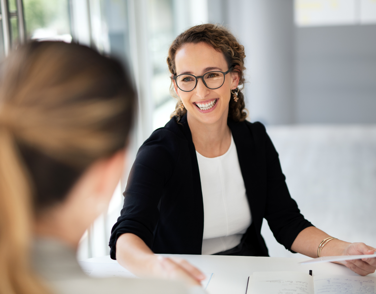 Zwei Frauen unterhalten sich lächelnd an einem Bürotisch und tauschen Dokumente aus.