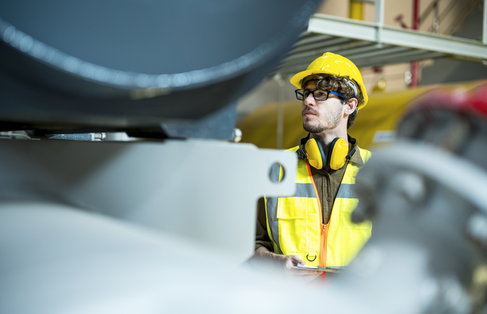 Fachkraft für Arbeitssicherheit mit Schutzhelm und Weste inspiziert Maschinen in einer Fabrikhalle.