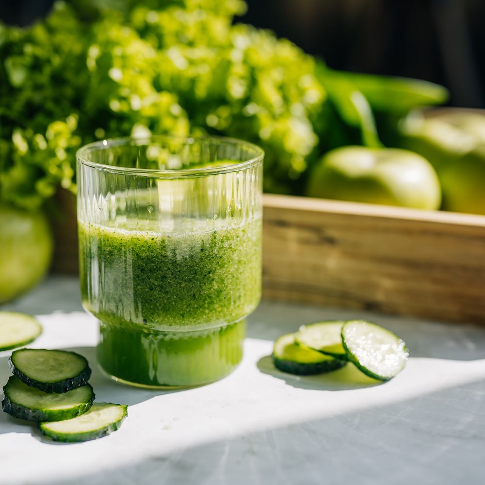 Ein Glas grüner Smoothie mit Gurkenscheiben auf einem Tisch, im Hintergrund Salat und Äpfel.