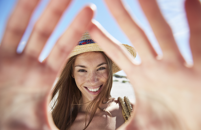 Junge Frau mit Strohhut und lachendem Gesicht, Hände formen einen Rahmen, blauer Himmel im Hintergrund.