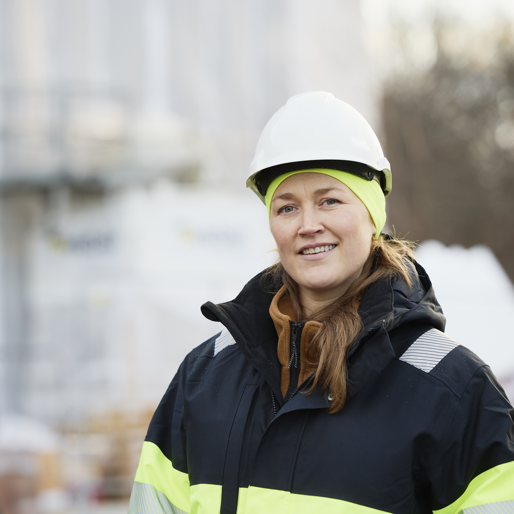 Frau in Schutzkleidung und Helm auf einer Baustelle lächelt in die Kamera.