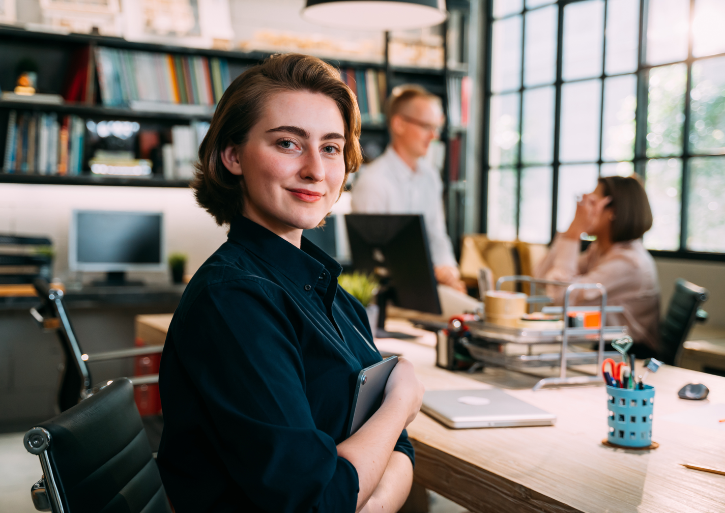 Frau sitzt im Büro, hält ein Tablet und blickt in die Kamera, im Hintergrund unterhalten sich Kollegen.