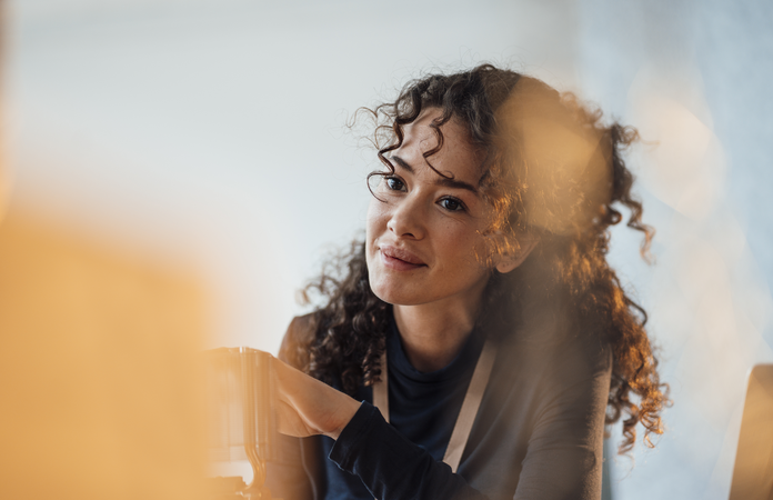 Eine junge Frau mit lockigem Haar sitzt an einem Tisch und lächelt freundlich.