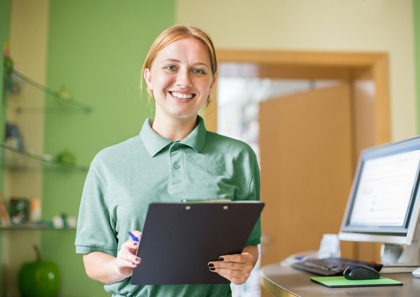 Eine Frau im grünen Polo-Shirt steht hinter dem Empfang einer Arztpraxis, lächelnd mit Klemmbrett in der Hand.