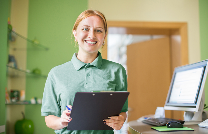 Eine Frau im grünen Polo-Shirt steht hinter dem Empfang einer Arztpraxis, lächelnd mit Klemmbrett in der Hand.