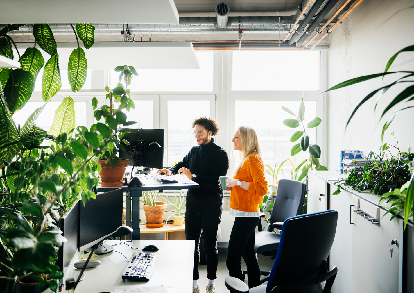 Zwei Personen arbeiten in einem modernen Büro voller Pflanzen, eine steht an einem Stehpult.