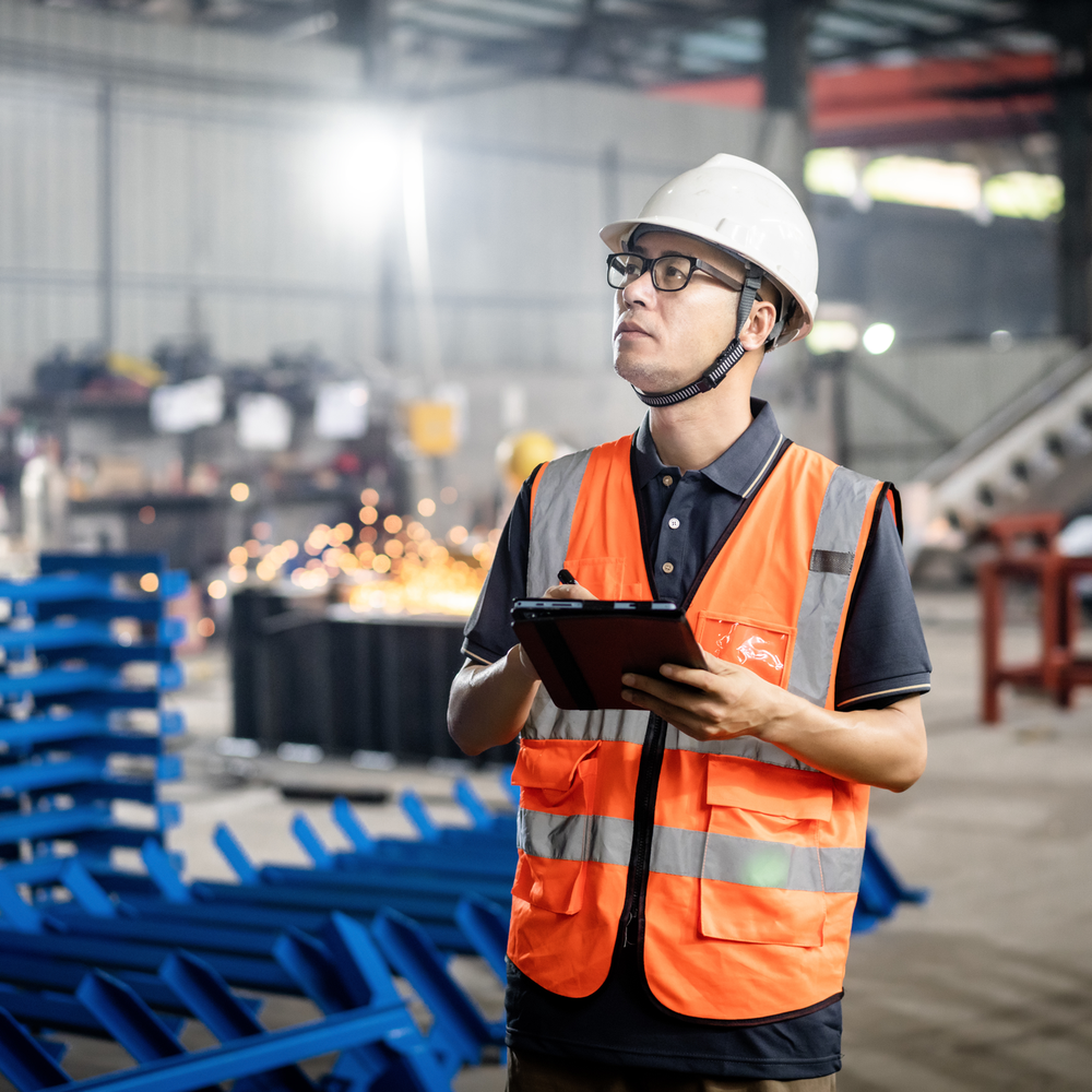 Ein Arbeiter mit Schutzweste und Helm überprüft Maschinenteile in einer Produktionshalle.
