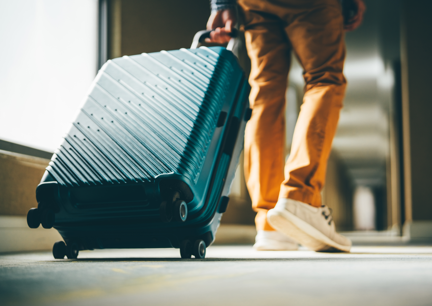 erson in Jeans und Turnschuhen zieht blauen Koffer ueber Hotelflur