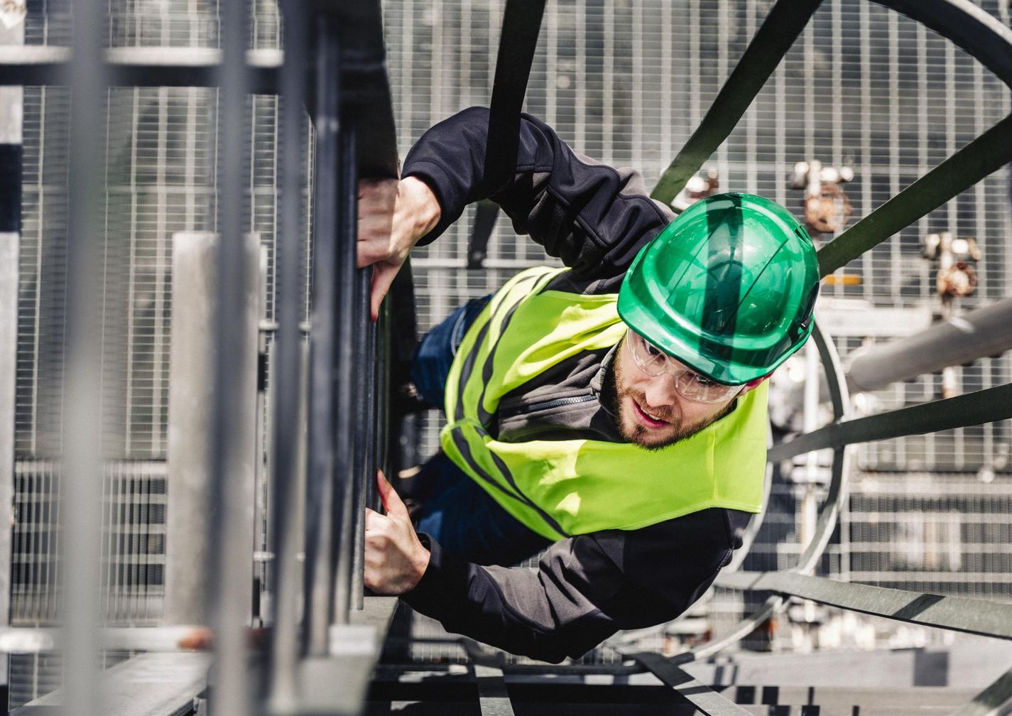 Worker wearing a green hard hat and safety vest climbs an industrial metal ladder on a construction site, looking upward.