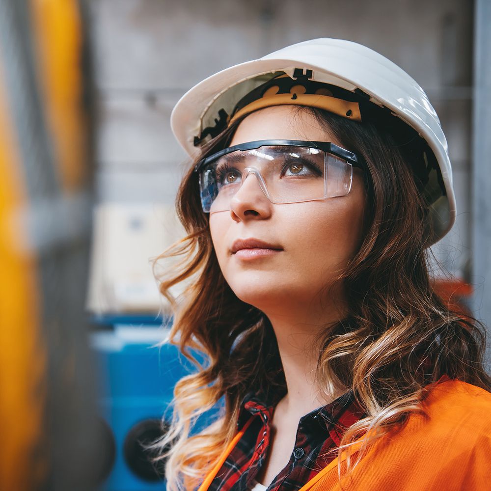 ngenieurin mit Schutzhelm und Brille inspiziert eine Maschine in einer Fabrikhalle.