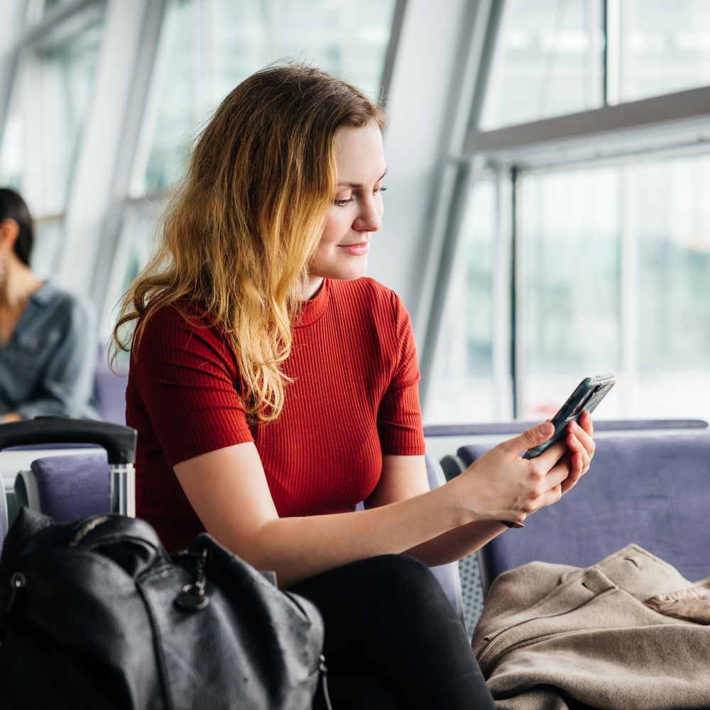 Frau in rotem Shirt sitzt am Flughafen und schaut auf ihr Smartphone, mit Gepäck neben sich.