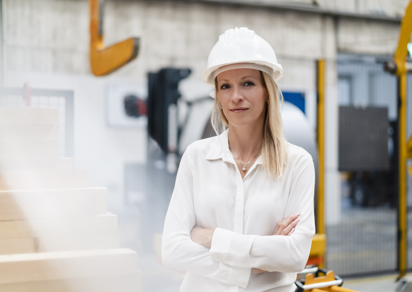 Woman in a white shirt and hard hat stands confidently in an industrial setting with machinery in the background.