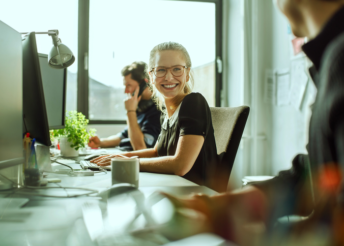 rei Personen arbeiten gemeinsam an Computern in einem hellen Büro und lachen dabei.
