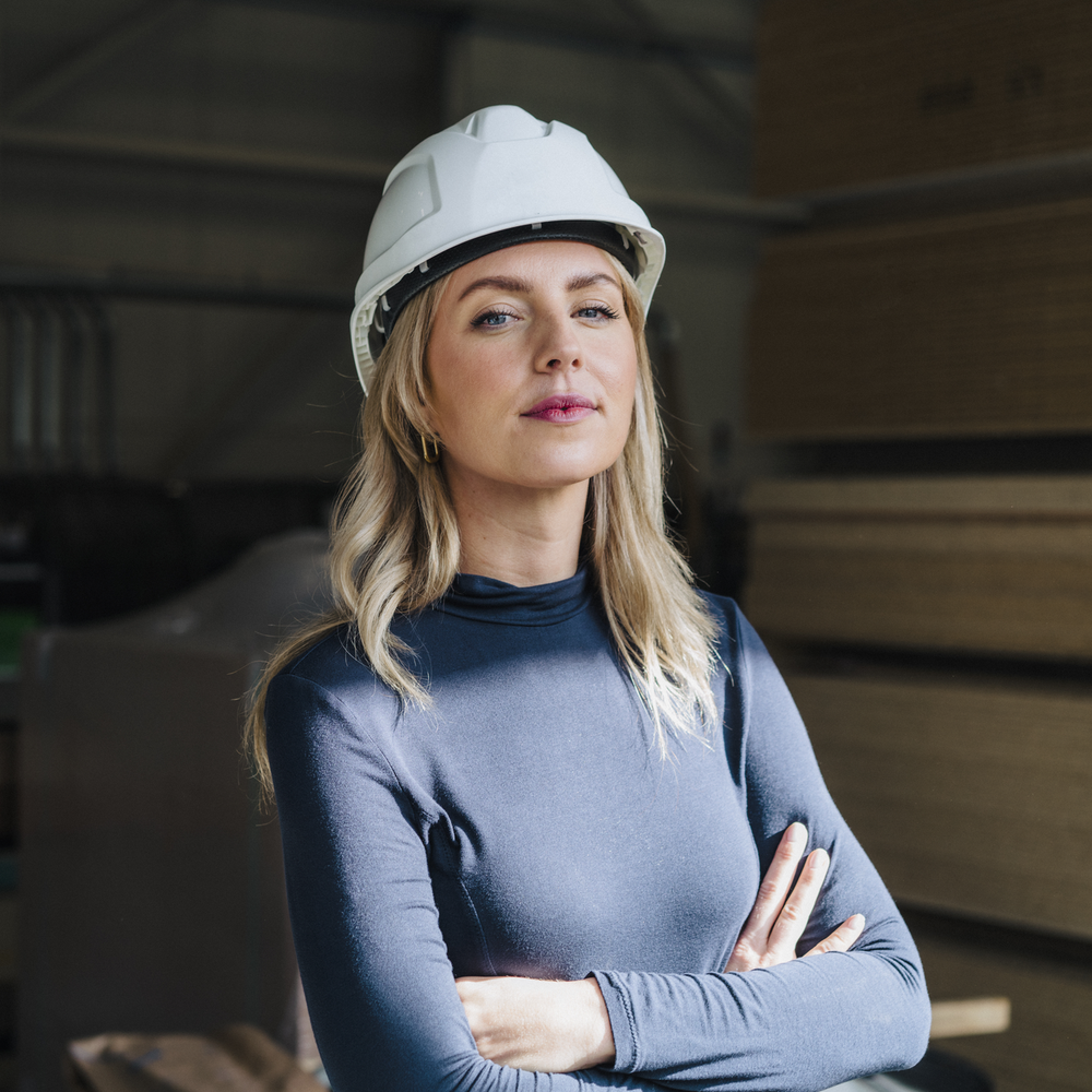 Frau mit Schutzhelm und verschraenkten Armen in einer Fabrikhalle