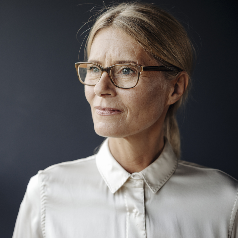 Portrait einer Frau mit Brille, die nachdenklich aus einem Fenster schaut.