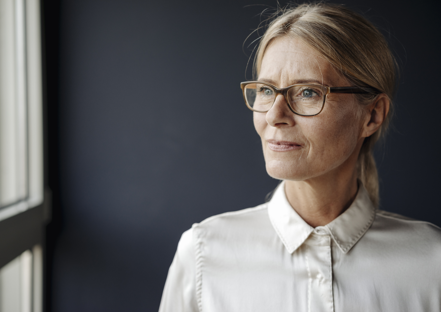 Portrait einer Frau mit Brille, die nachdenklich aus einem Fenster schaut.