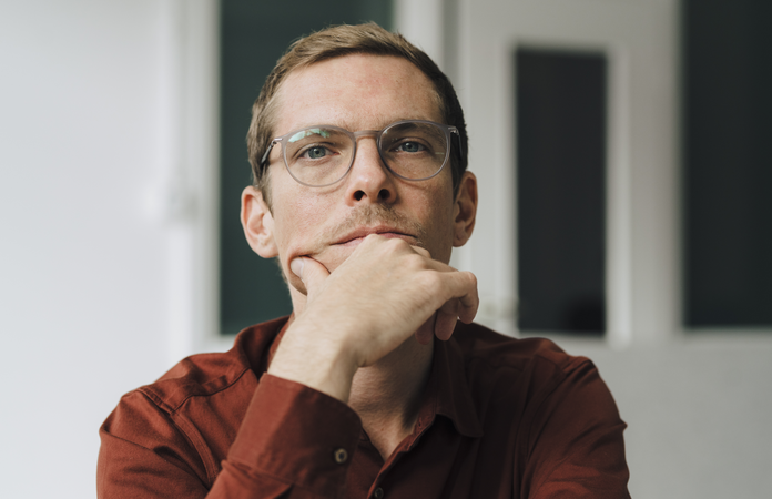 Nachdenklicher Mann mit Brille und rotem Hemd stützt das Kinn mit der Hand ab und blickt konzentriert nach vorn.