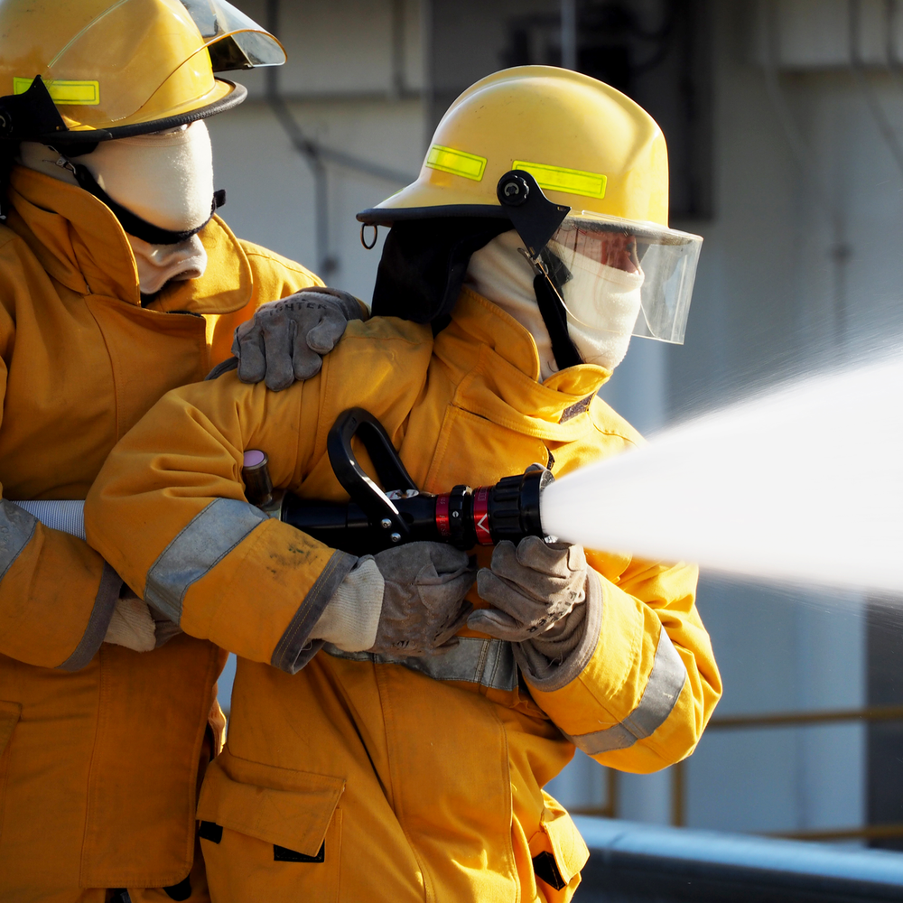 Zwei Feuerwehrleute in voller Schutzkleidung spritzen Wasser aus einem Schlauch waehrend einer Uebung.