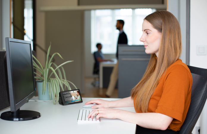 Eine Frau sitzt am Schreibtisch vor Ihrem Arbeitsplatz, KICO steht im Hintergrund