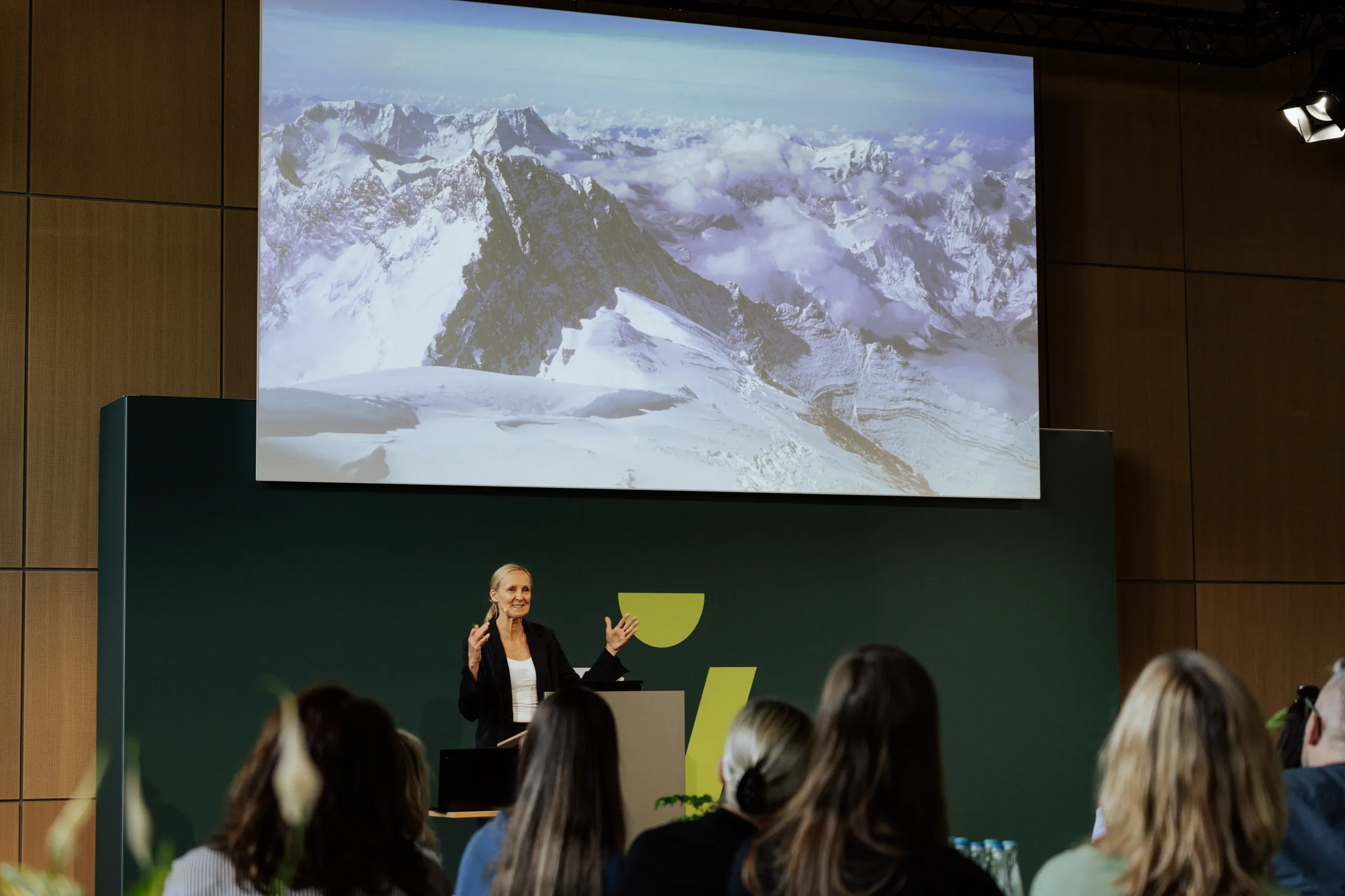 A woman in a black blazer presents to an audience, standing in front of a screen displaying snowy mountain peaks.