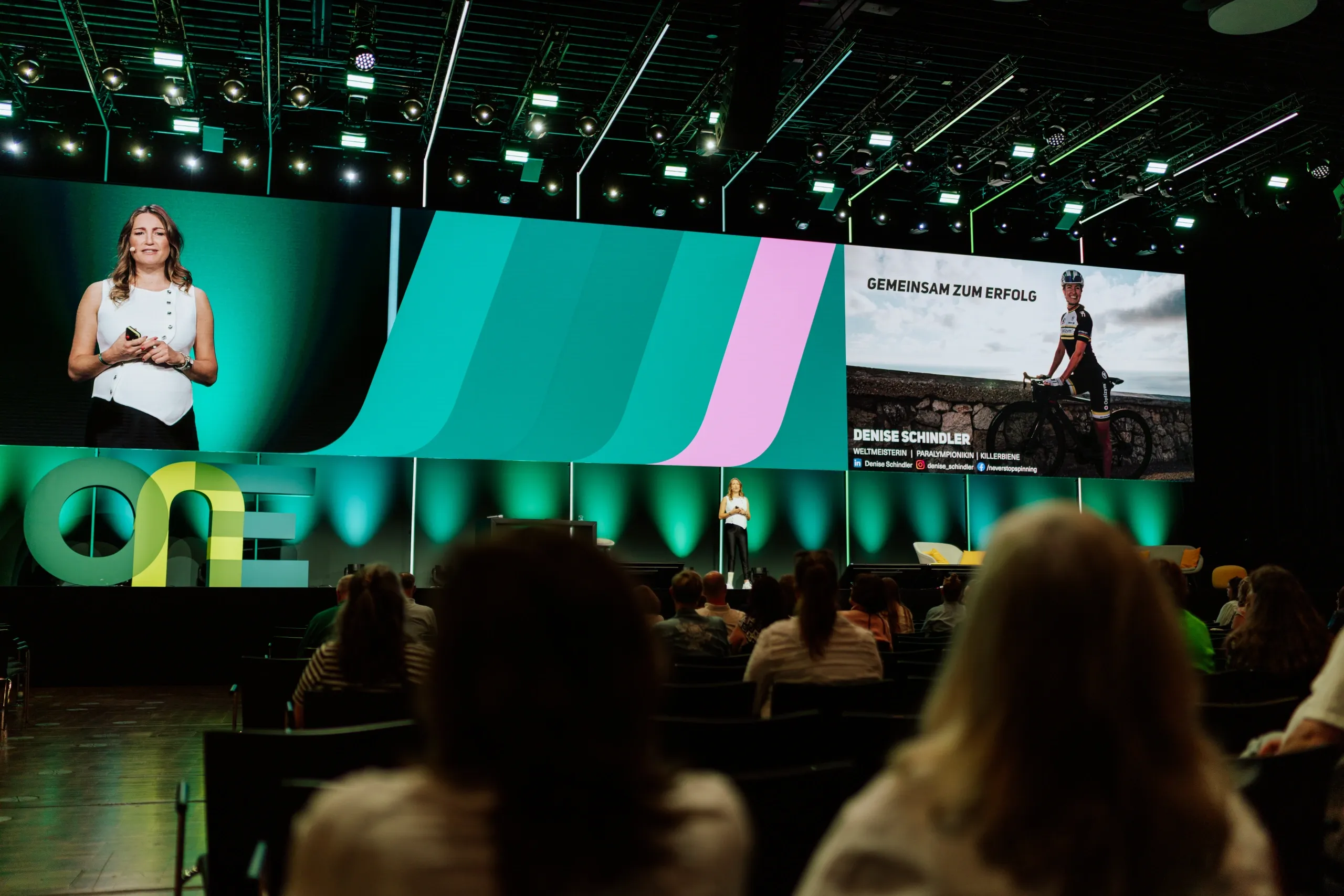A woman speaks on stage at a conference with a large screen displaying graphics and text. Audience members are seated in the foreground.