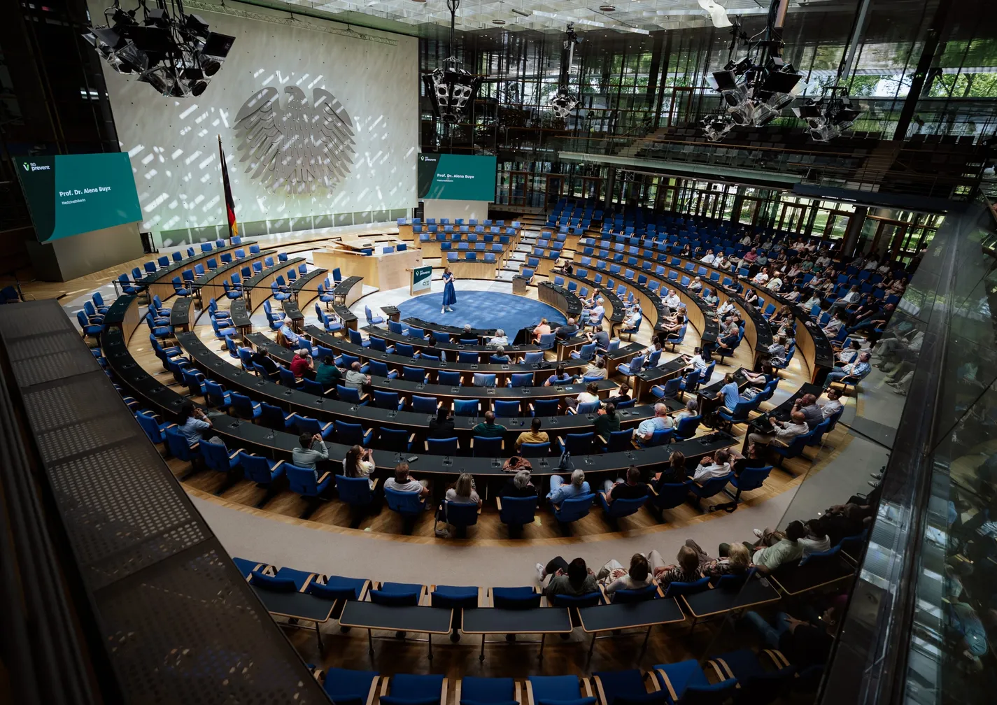 Aerial view of a circular auditorium with people seated, a speaker on stage, and a large emblem on the wall under bright lights.