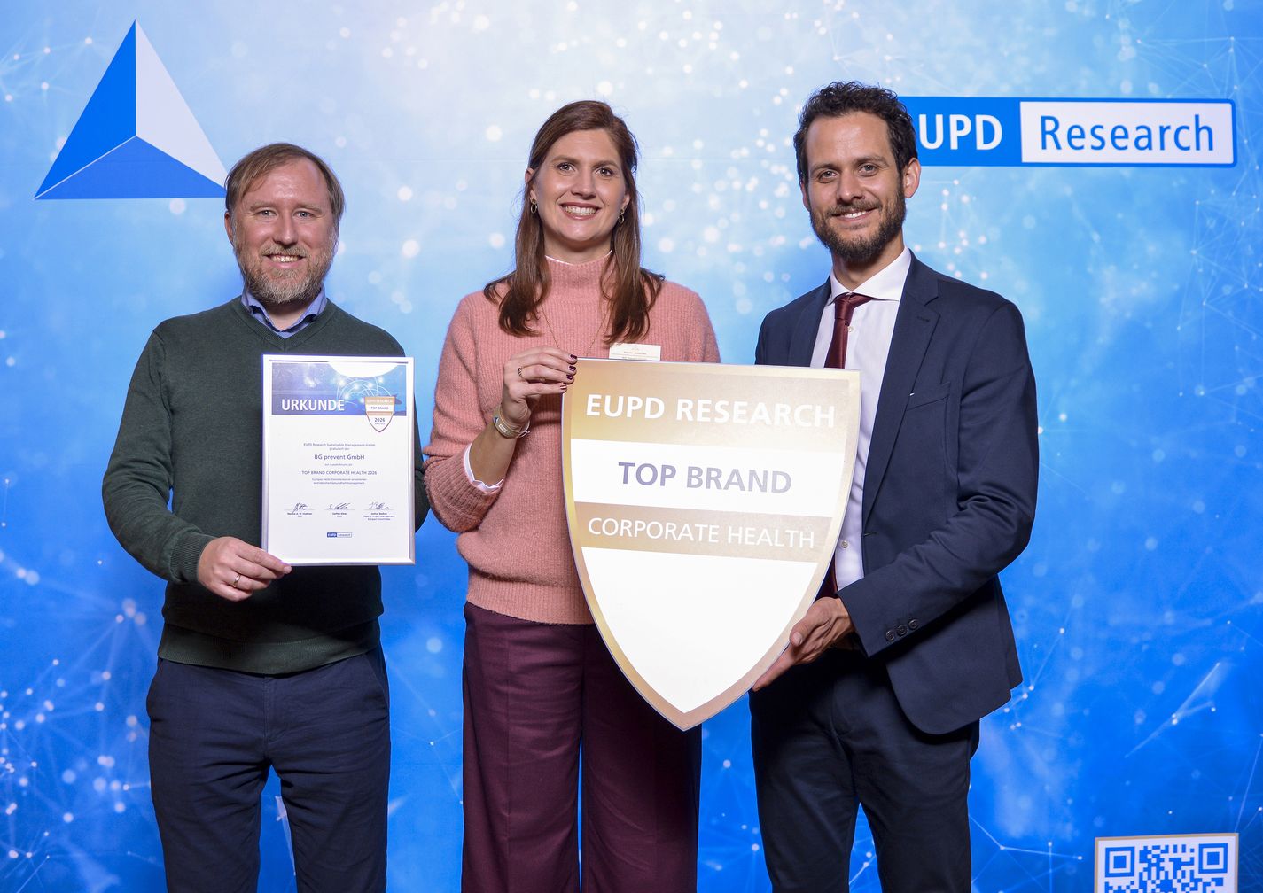 Vor einem blauen Hintergrund stehen drei Personen. Links en Mann mit grünem Pullover und blauer Hose, in der Hand eine Urkunde. In der Mitte eine pinkfarben gekleidete Frau, die mit dem Mann im blauen Anzug, rechts von ihr ein Siegel hält. Alle drei lächeln.