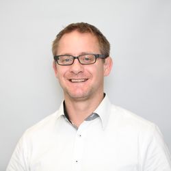 Smiling person with glasses, wearing a white shirt, against a light gray background.