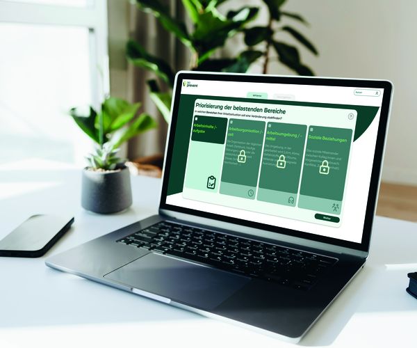 Laptop auf Schreibtisch zeigt die BG prevent-Webanwendung pria psych, daneben Smartphone und Pflanze.
