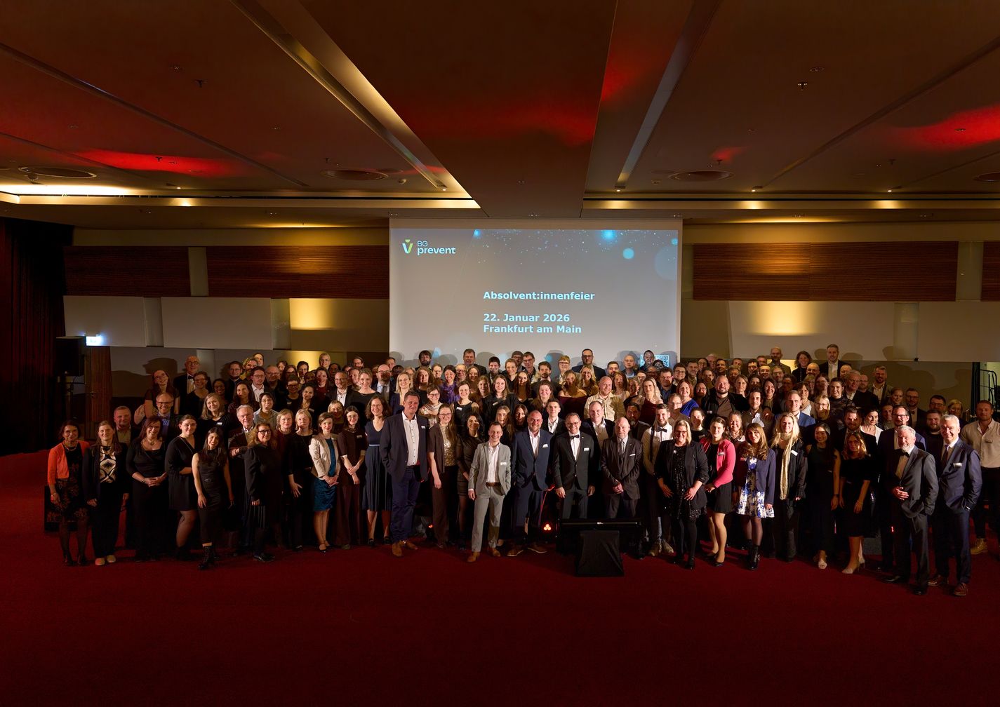 Large group of people posing in a banquet hall, with a presentation screen displaying "Absolventenfeier" and the date January 22, 2016.