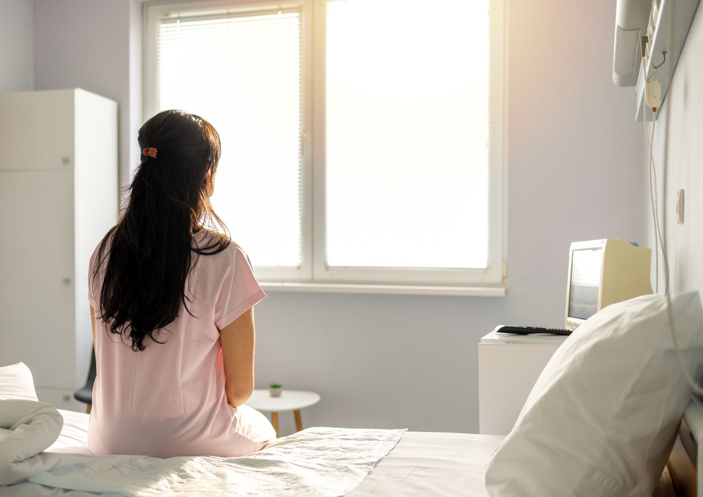 Eine Frau sitzt mit dem Rücken zugewandt auf einem Bett und schaut Richtung Fenster