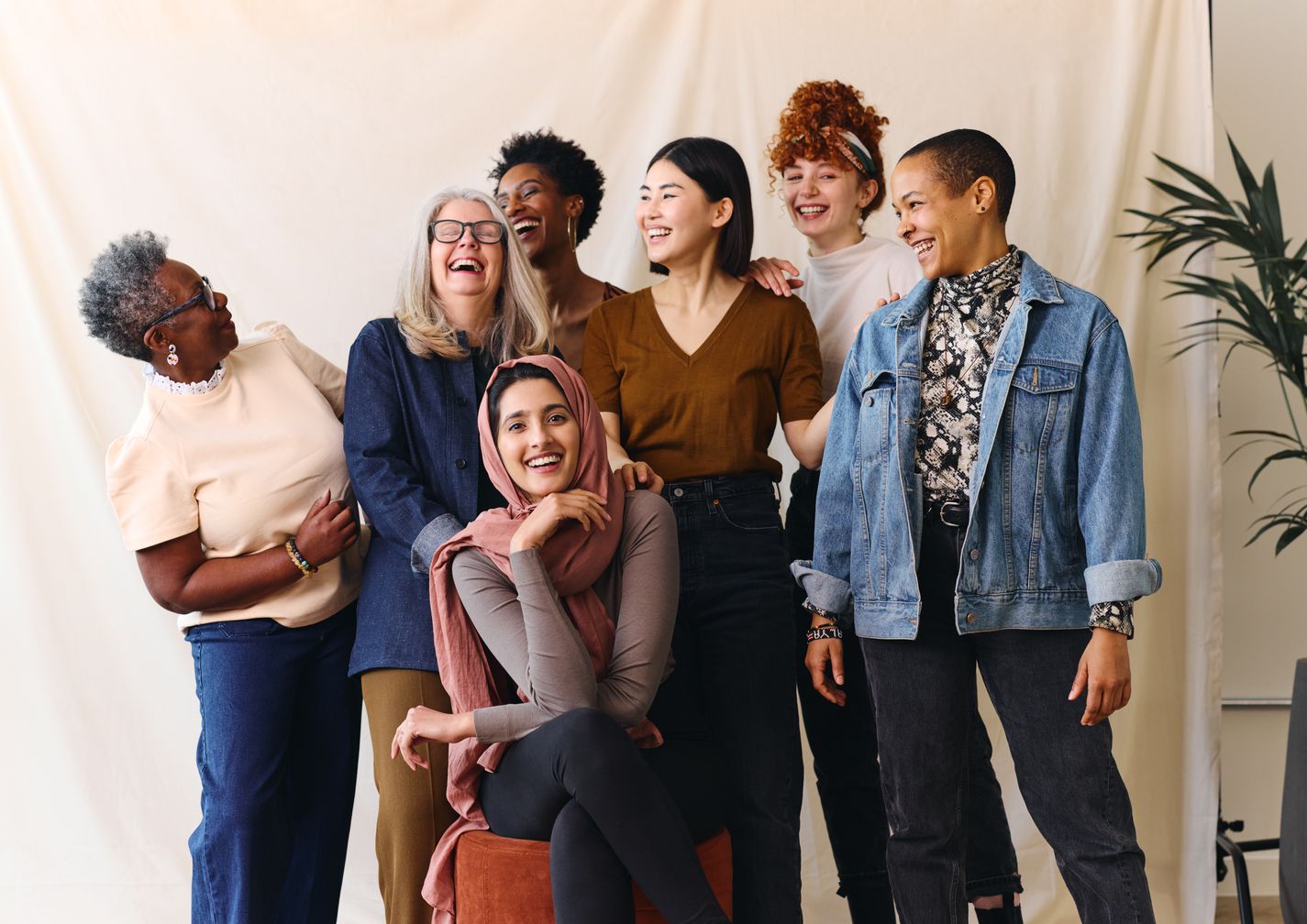 Eine diverse Gruppe von sieben Frauen unterschiedlichen Alters und mit verschiedenen ethnischen Hintergründen steht und sitzt zusammen vor einem neutralen Hintergrund. Sie lachen und schauen sich gegenseitig an, wodurch eine freundliche und entspannte Stimmung entsteht. Eine Frau sitzt vorne auf einem Hocker und trägt ein rosa Kopftuch, während die anderen hinter ihr stehen und lässige Kleidung wie Jeansjacke, Pullover und Blusen tragen. Eine Zimmerpflanze ist am rechten Bildrand zu sehen.
