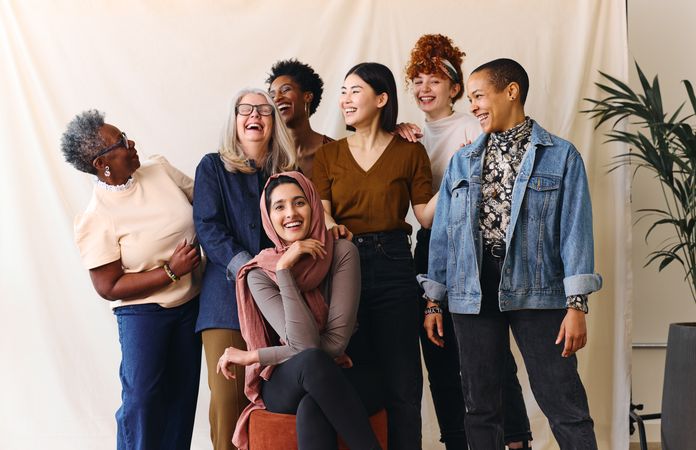 Eine diverse Gruppe von sieben Frauen unterschiedlichen Alters und mit verschiedenen ethnischen Hintergründen steht und sitzt zusammen vor einem neutralen Hintergrund. Sie lachen und schauen sich gegenseitig an, wodurch eine freundliche und entspannte Stimmung entsteht. Eine Frau sitzt vorne auf einem Hocker und trägt ein rosa Kopftuch, während die anderen hinter ihr stehen und lässige Kleidung wie Jeansjacke, Pullover und Blusen tragen. Eine Zimmerpflanze ist am rechten Bildrand zu sehen.