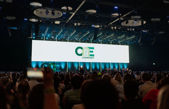 Bühnenbild auf dem One Company steht im Hintergrund, davor sitzen viele Menschen, deren Rücken man sieht
