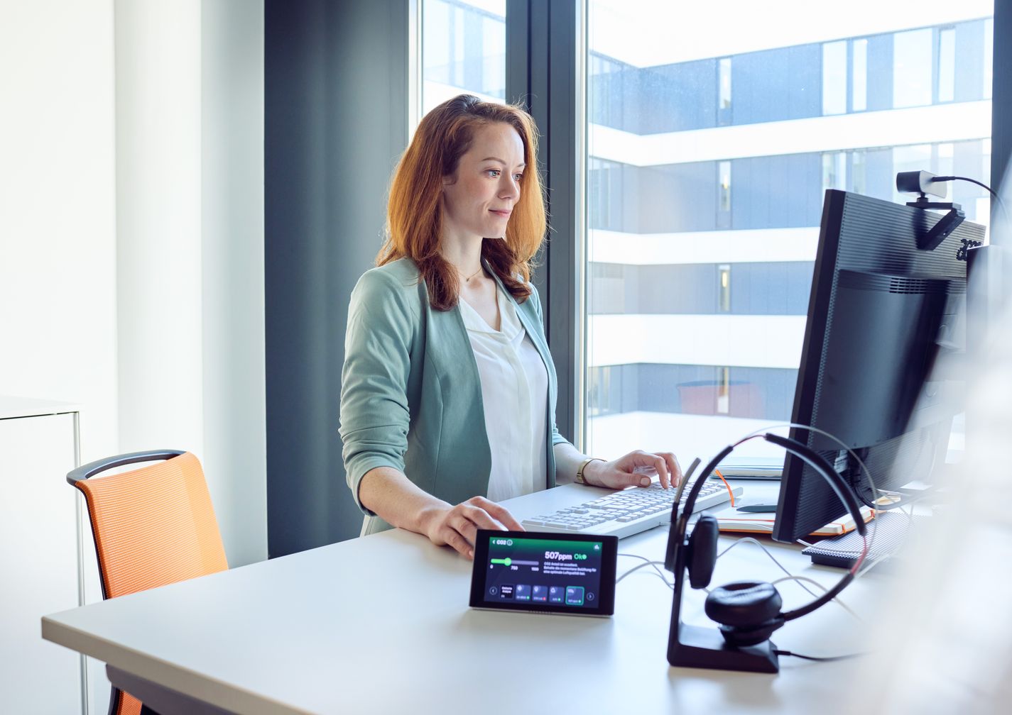 Frau arbeitet im Stehen an einem Computerarbeitsplatz mit Headset und KICO Gesundheitscoach auf dem Schreibtisch, modernes Bürogebäude im Hintergrund.