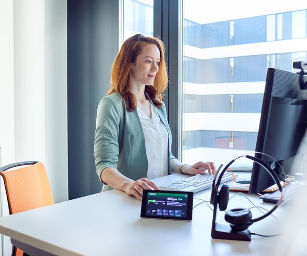 Frau arbeitet im Stehen an einem Computerarbeitsplatz mit Headset und KICO Gesundheitscoach auf dem Schreibtisch, modernes Bürogebäude im Hintergrund.