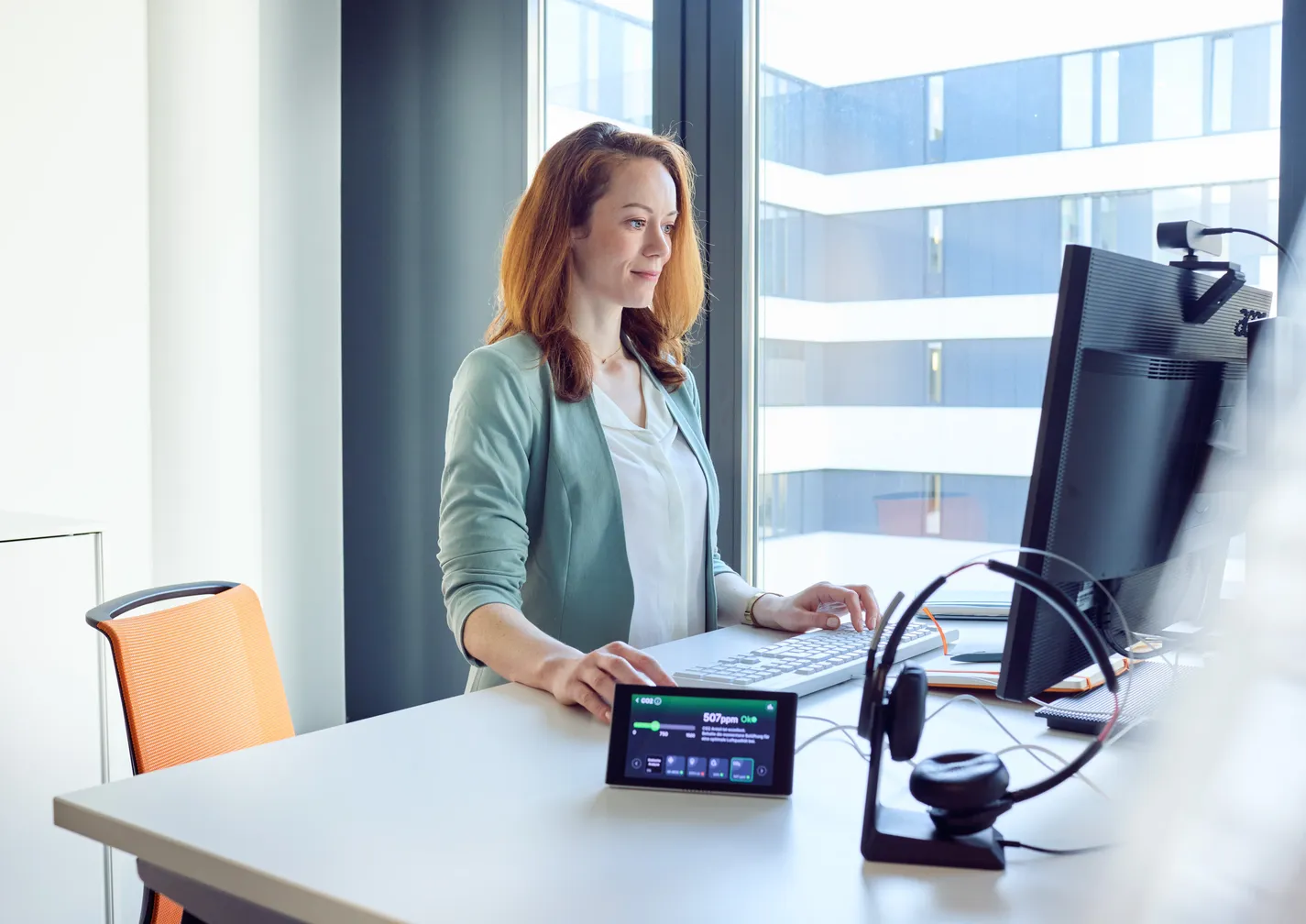 Frau arbeitet konzentriert im modernen Büro am Stehschreibtisch mit Blick auf den Monitor vor ihr und KICO – KI-Gesundheitscoach auf dem Tisch.