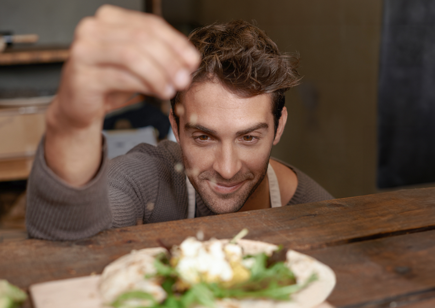 Man in a cozy setting garnishing a dish with herbs, smiling, focusing on the food placed on a wooden surface.