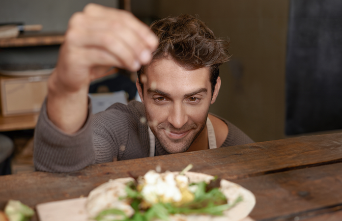 Man in a cozy setting garnishing a dish with herbs, smiling, focusing on the food placed on a wooden surface.