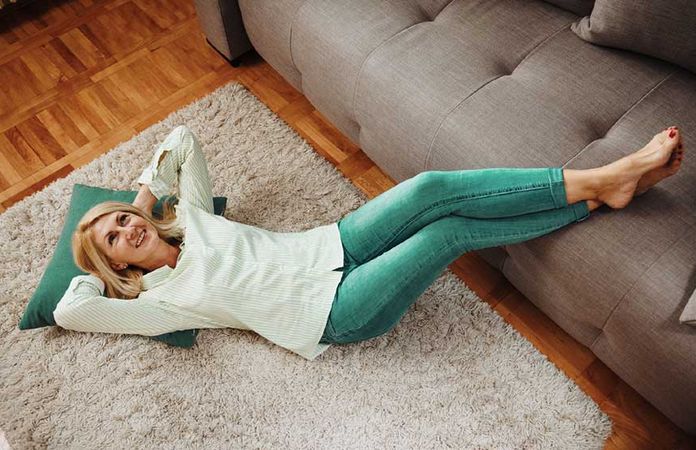 Eine Frau liegt entspannt auf dem Boden, die Beine auf dem Sofa und unter dem Kopf ein Kissen